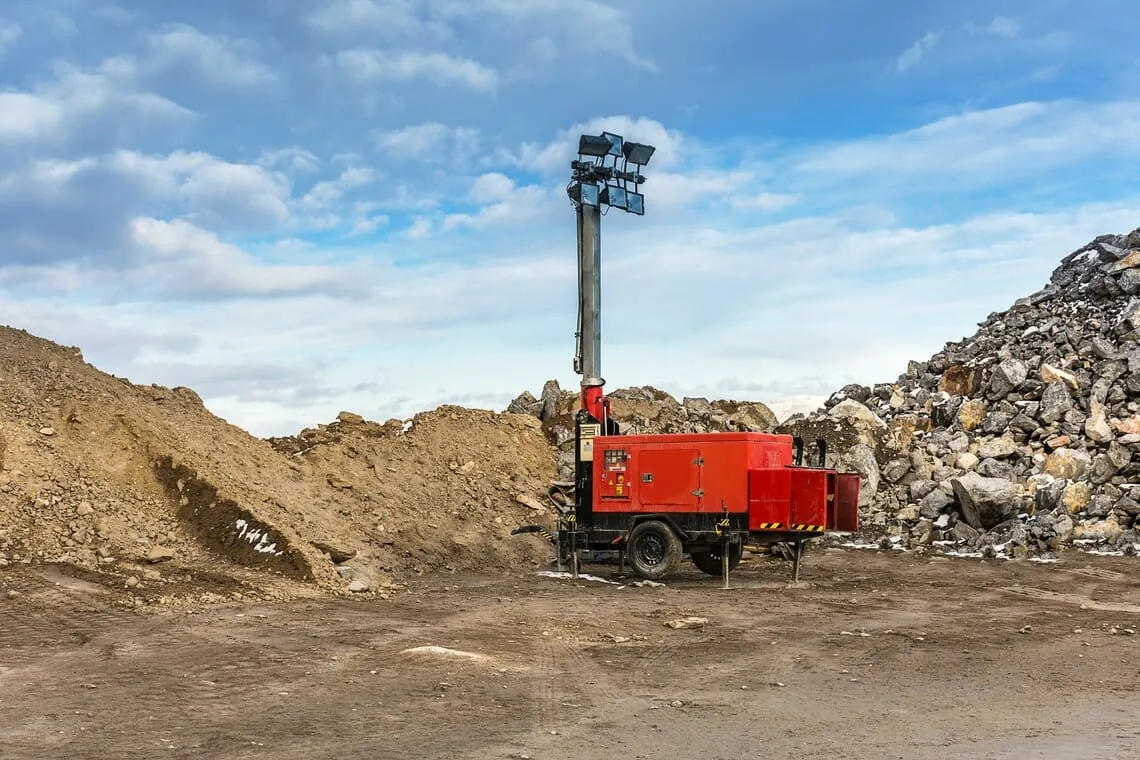 Mobiler Lichtmast mit Stromerzeuger und vier Flutlichtstrahlern auf einer Baustelle zwischen Erd- und Schotterhaufen